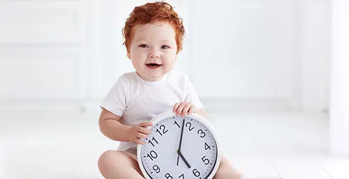 Nauka zegara - jak nauczyć dzieci odczytywać godzinę na zegarku? Mały rudy chłopczyk trzyma w rączkach zegar wskazówkowy.
