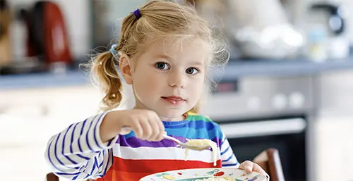 Probiotyk dla dzieci - dlaczego warto go stosować? Blondwłosa mała dziewczynka je owsiankę na śniadanie.
