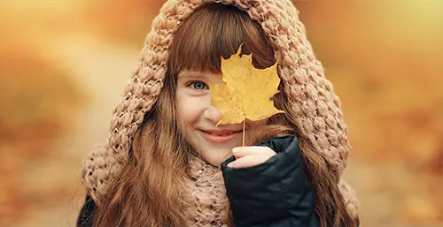 Witamina D dla dzieci – kiedy warto ją suplementować? Brązowowłosa dziewczynka uśmiecha się do aparatu zakrywając jedno oko jesiennym liściem.