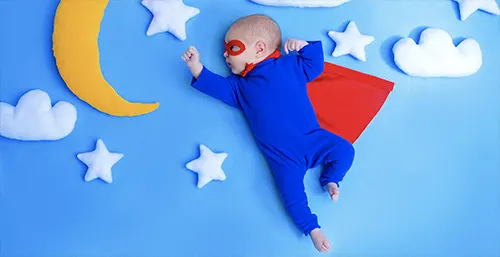 Witamina D dla niemowląt i noworodków - dlaczego warto ją stosować? Niemowlę przebrane za supermana śpi w otoczeniu pluszowego księżyca, gwiazdek i chmurek.