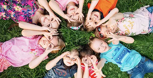 Witaminy na odporność dla dzieci. Dzieci leżą na zielonej trawie w kółku i przytykają palce do oczu jakby trzymały lornetkę.