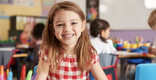 Lęk przed szkołą - jak przezwyciężyć fobię szkolną? Dziewczynka w klasie uśmiecha się do aparatu.
