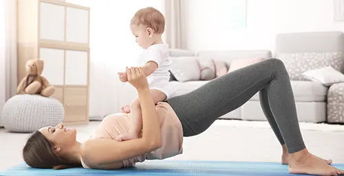 Brzuch po porodzie - jak odzyskać sylwetkę sprzed ciąży? Mama w stroju sportowym leży na plecach na macie w salonie i podnosi na biodrach niemowlę.