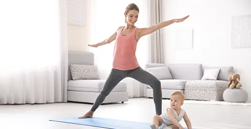 Ćwiczenia po porodzie - jak ćwiczyć po cesarce, a jak po porodzie naturalnym? Kobieta w legginsach i sportowej koszulce ćwiczy jogę na macie w salonie, na której siedzi też niemowlę.