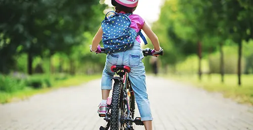 Jak zachęcić dziecko do aktywności fizycznej? Ćwiczenia i sporty dla dzieci. Dziewczynka z plecakiem siedzi na rowerze, odwrócona tyłem do aparatu.