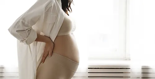 Poród naturalny a cesarskie cięcie - co warto wiedzieć? Kobieta w średniozaawansowanej ciąży w cielistej zabudowanej bieliźnie i rozpiętej białej koszuli stoi przy oknie na tle białej firanki.
