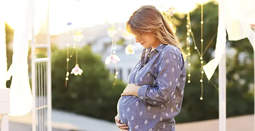 Witamina D w ciąży - dlaczego warto ją suplementować? Kobieta w ciąży w niebieskiej koszuli w ogrodzie.