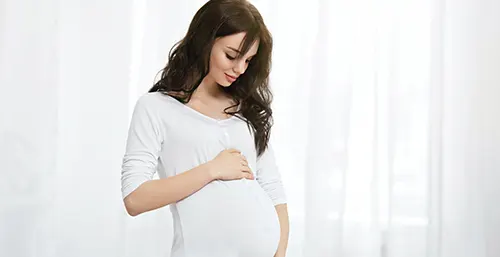 Zaparcia w ciąży - jakie są najlepsze sposoby i jak sobie z nimi radzić? Kobieta w zaawansowanej ciąży w białej sukience trzyma się za brzuch.