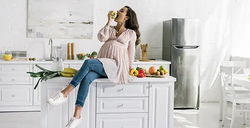 Zgaga w ciąży - jak sobie z nią radzić? Domowe sposoby na zgagę. Kobieta w ciąży je jabłko, siedząc na blacie w kuchni.